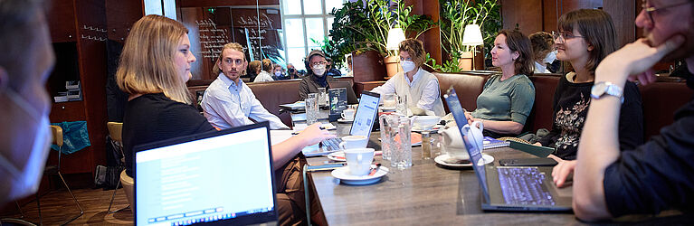 Eine Gruppe von Personen sitzt in einem Café an einem großen Tisch und sprechen miteinander. Notebooks und Arbeitsmaterial befinden sich auf dem Tisch. 