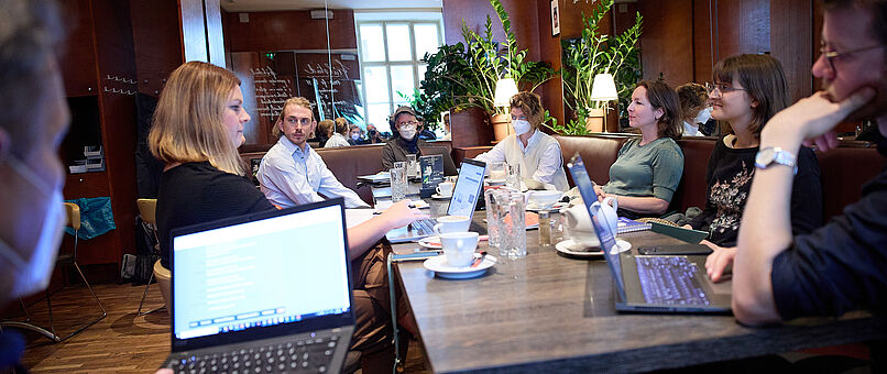 Eine Gruppe von Personen sitzt in einem Café an einem großen Tisch und sprechen miteinander. Notebooks und Arbeitsmaterial befinden sich auf dem Tisch. 